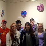 A group of 8 students stand in front of a wall featuring painted and decorated wooden hearts