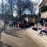 A crowd gathers and looks on as performers stand on the steps of Gorgas Library